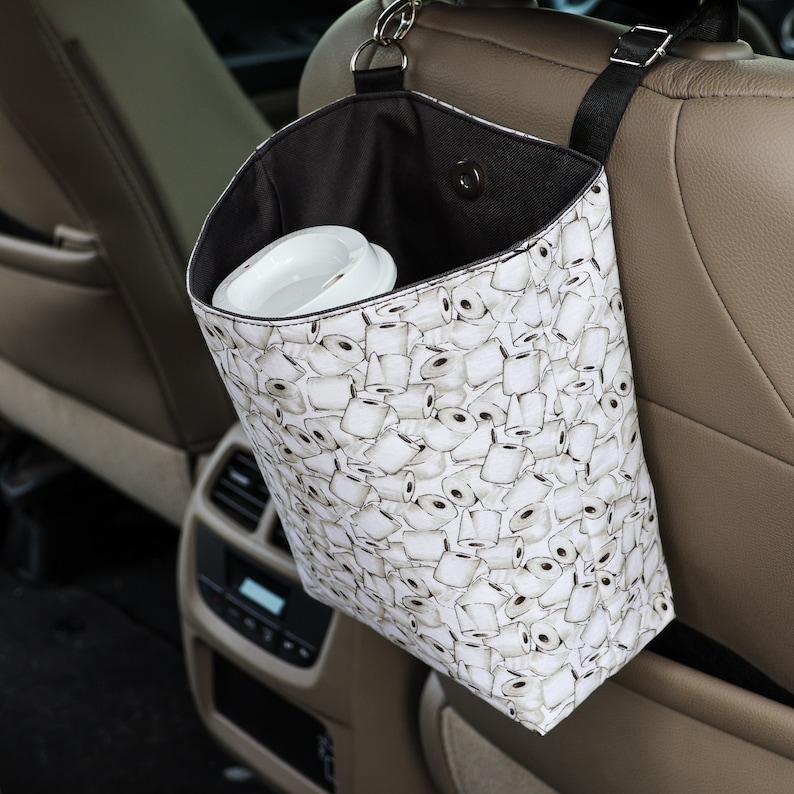 Peut inclure: Un sac poubelle de voiture blanc avec un dessus noir et une sangle noire. Le sac est suspendu au dossier d'un si&egrave;ge de voiture. Le sac est recouvert d'un motif de rouleaux de papier toilette.