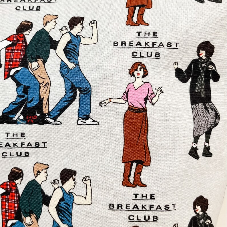 May include: A white fabric with a repeating pattern of characters from the movie The Breakfast Club. The characters are illustrated in a cartoon style and are wearing their iconic outfits from the film. The text "The Breakfast Club" is repeated throughout the fabric.