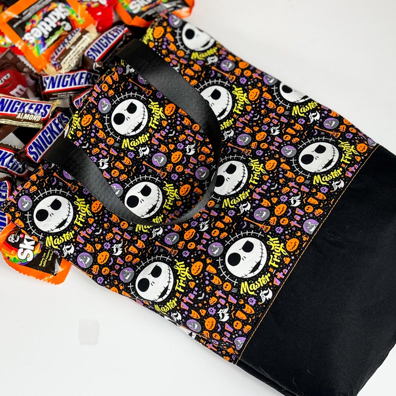 Puede incluir: Bolsa de tela negra y blanca con tem&aacute;tica de Halloween que presenta un patr&oacute;n repetido de la cara de Jack Skellington y las palabras "Master Fright". La bolsa tiene asas negras y est&aacute; hecha de una tela resistente.