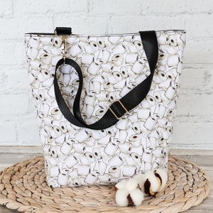 Peut inclure: Un sac fourre-tout blanc avec une bandouli&egrave;re noire et un motif de rouleaux de papier toilette blancs sur fond blanc. Le sac est pos&eacute; sur une surface en paille tress&eacute;e.