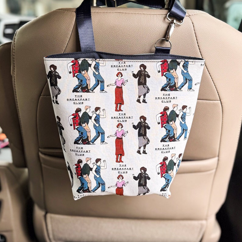 May include: A white tote bag with a black handle and a repeating pattern of characters from the movie "The Breakfast Club". The characters are depicted in a cartoon style and are dancing. The text "The Breakfast Club" is repeated throughout the pattern.