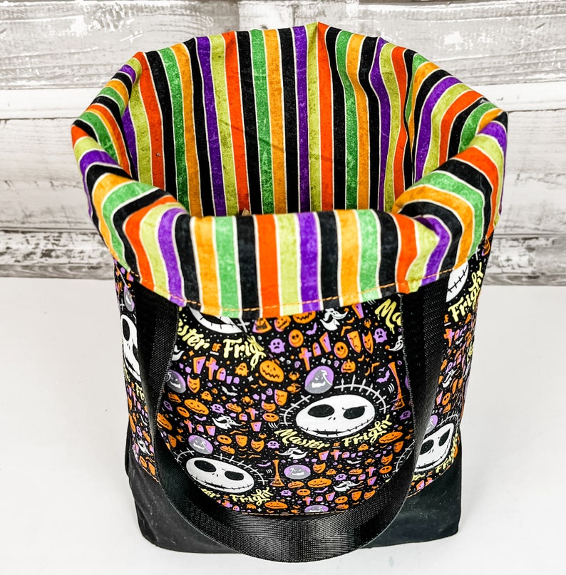 Puede incluir: Una bolsa de tela negra y naranja con tem&aacute;tica de Halloween con un forro de tela a rayas. La bolsa presenta un patr&oacute;n repetido de personajes de Jack Skellington y las palabras "Master of Fright".