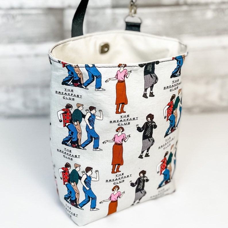 May include: White canvas tote bag with a black handle and a colorful print of characters from the movie "The Breakfast Club". The print features the characters in various poses and includes the movie title.