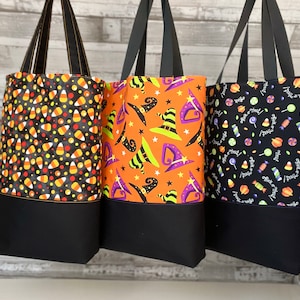 Puede incluir: Tres bolsas de tela con tem&aacute;tica de Halloween. Una bolsa presenta un patr&oacute;n de ma&iacute;z dulce negro y naranja, otra tiene un patr&oacute;n de sombrero de bruja negro y naranja, y la tercera tiene un patr&oacute;n de caramelo negro y blanco con el texto "Trick or Treat".