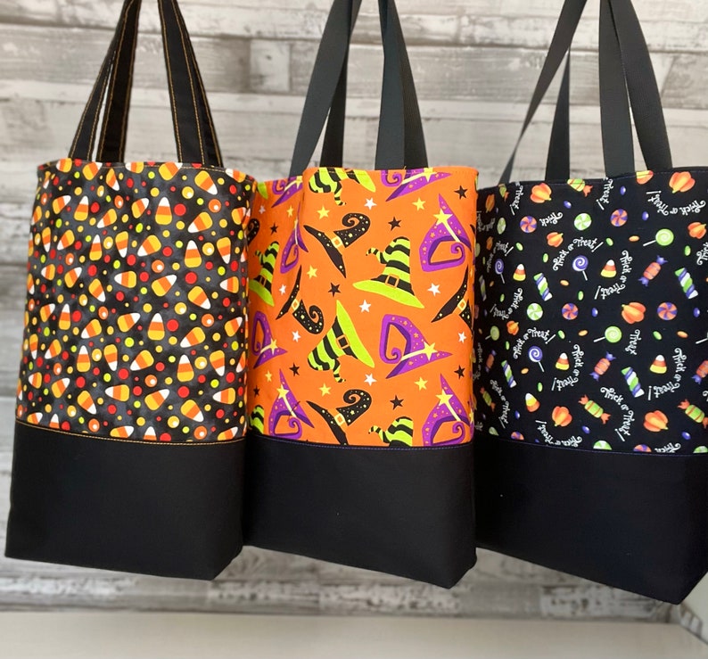Puede incluir: Tres bolsas de tela con tem&aacute;tica de Halloween. Una bolsa presenta un patr&oacute;n de ma&iacute;z dulce negro y naranja, otra tiene un patr&oacute;n de sombrero de bruja negro y naranja, y la tercera tiene un patr&oacute;n de caramelo negro y blanco con el texto "Trick or Treat".