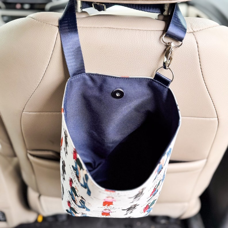 May include: A white fabric car trash bag with a blue lining and a black strap. The bag is hanging from the headrest of a car seat. The bag has a cartoon design of people walking.