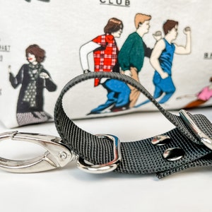 May include: A black adjustable strap with a silver hook and a silver buckle. The strap is attached to a white fabric bag with a print of characters from the movie "The Breakfast Club".