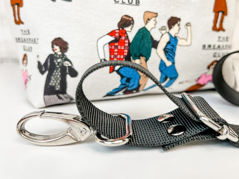 May include: A black adjustable strap with a silver hook and a silver buckle. The strap is attached to a white fabric bag with a print of characters from the movie "The Breakfast Club".