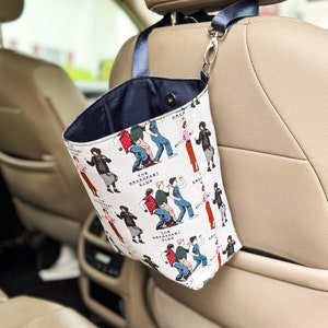 May include: A white fabric car trash can with a blue drawstring and a black handle. The can is decorated with a repeating pattern of characters from the movie "The Breakfast Club" and the text "The Breakfast Club".