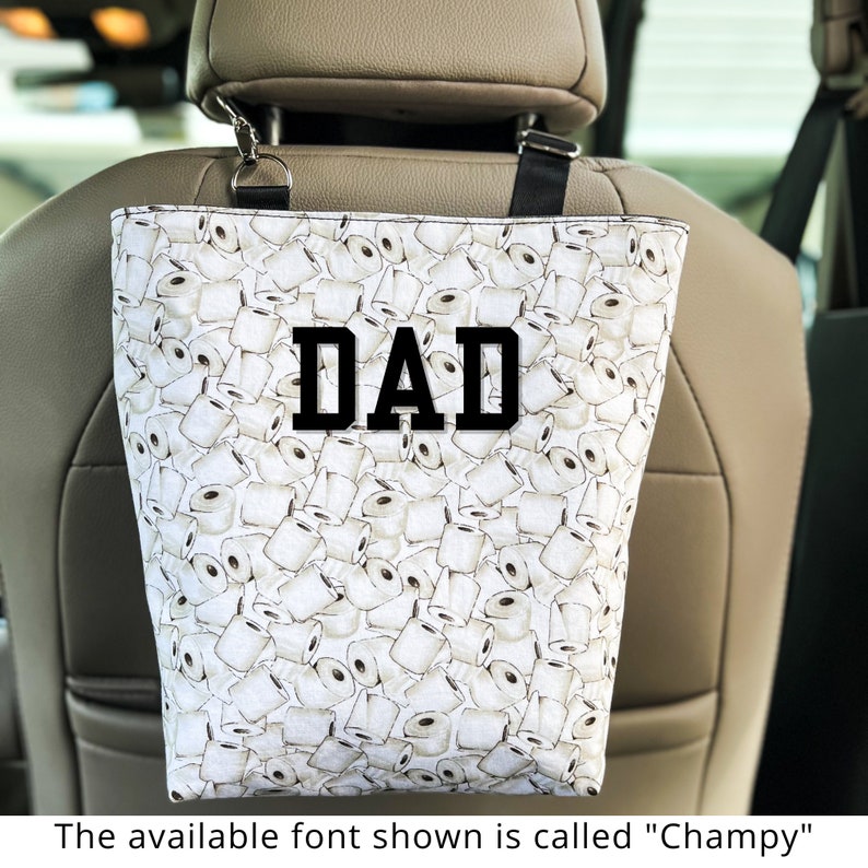 Peut inclure: Sac fourre-tout blanc avec une sangle noire et un motif de rouleaux de papier toilette. Le sac porte l'inscription "DAD" en noir sur le devant.