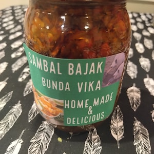 May include: A glass jar filled with a red and orange sauce labelled "Sambal Bajak Bunda Vika Home & Delicious".