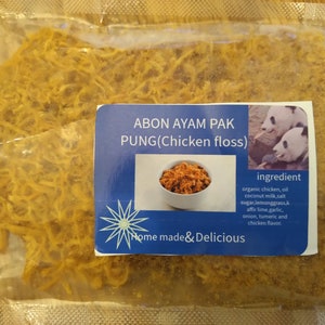 May include: A clear plastic bag filled with golden-brown chicken floss. A blue label displays the text "ABON AYAM PAK PUNG (Chicken floss)" and a small image of the product. The label also lists ingredients and features a panda image.