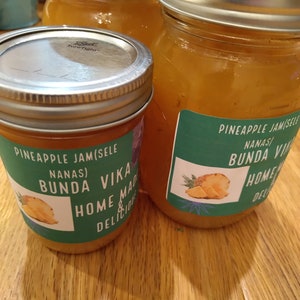 May include: Three glass jars filled with a light orange liquid. The jars have silver lids and green labels with the text "Pineapple Jam (Sele Nana's) Bunda Vika Home Made Delicious" and a picture of a pineapple.