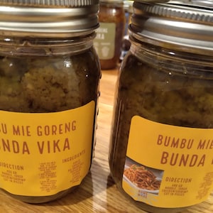 Puede incluir: Dos tarros de vidrio llenos de una pasta verde etiquetada "BUMBU MIE GORENG BUNDA VIKA" con fondo amarillo y texto negro. Los tarros tienen tapas plateadas y están sobre una superficie de madera. Las etiquetas incluyen instrucciones e ingredientes para hacer un plato.