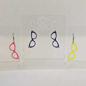 Könnte beinhalten: Drei Paar Hängeohrringe mit einer Brillenform. Die Ohrringe sind in Rot, Blau und Gelb.