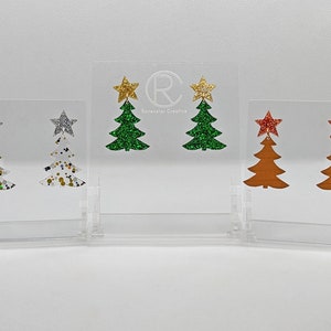 Può includere: Tre paia di orecchini a forma di albero di Natale. Il primo paio è bianco con glitter dorati e blu, il secondo paio è verde con glitter dorati e il terzo paio è marrone con glitter rossi.