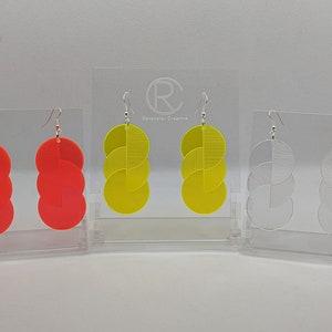 May include: Three pairs of geometric earrings with a layered circle design. The earrings are displayed on a white stand. The earrings are available in red, yellow, and clear.