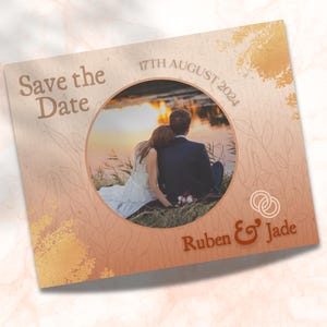 May include: Save the Date card with a photo of a couple in front of a lake at sunset. The card is decorated with a gold and brown watercolor background and the text "Save the Date" in a brown font. The date "17th August 2024" is printed in a brown font. The couple's names "Ruben & Jade" are printed in a brown font.