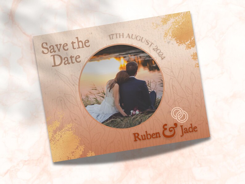 May include: Save the Date card with a photo of a couple in front of a lake at sunset. The card is decorated with a gold and brown watercolor background and the text "Save the Date" in a brown font. The date "17th August 2024" is printed in a brown font. The couple's names "Ruben & Jade" are printed in a brown font.