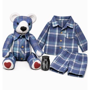 May include: A blue plaid teddy bear, shirt, and shorts set. The bear has white ears and paws, and red heart-shaped accents on its feet. A small black container with white text is also included.