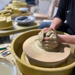 May include: A person's hands shaping a lump of clay on a pottery wheel. The wheel is yellow and has a white ceramic base. The clay is a light brown colour.
