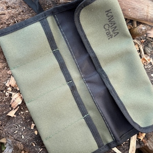 Puede incluir: Un estuche enrollable para herramientas de lona verde oliva con ribete negro y el texto "Kavana Craft" impreso en la solapa. El rollo está abierto, revelando el interior. El estuche enrollable para herramientas está sobre una superficie de madera desgastada.