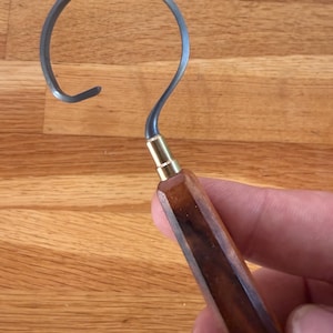 May include: A metal hook tool with a curved, question mark-shaped end. The tool has a dark wooden handle with a brass fitting. The hook is held against a wooden surface.