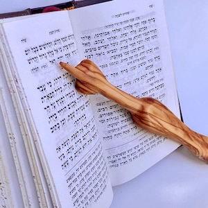 Może przedstawiać: Drewniany wskaźnik z końcówką w kształcie dłoni, leżący na otwartej książce z tekstem hebrajskim. Wskaźnik wykonany jest z drewna oliwnego i ma naturalny rysunek drewna.