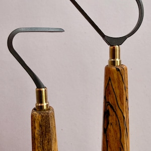 Può includere: Due strumenti per ceramica con manici in legno e lame in metallo. I manici sono marrone chiaro con venature nere. Le lame in metallo sono grigio scuro e attaccate ai manici con metallo color oro.