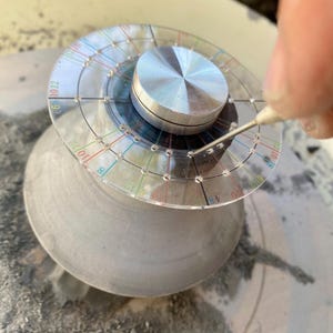 Könnte beinhalten: Ein klares, kreisförmiges Töpferwerkzeug mit einem silbernen Metallmittelteil und einem äußeren Ring aus klarem Kunststoff, der mit Zahlen und farbigen Linien markiert ist. Eine Hand verwendet ein Metallwerkzeug, um die graue Töpferscheibe zu markieren. Das Rad hat eine raue Textur.