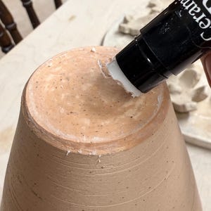 Puede incluir: Un recipiente de cerámica de color marrón claro, de forma cónica, con estrías horizontales visibles en su superficie. Un aplicador negro con el texto blanco "Cerami" aplica una sustancia blanca en el borde superior del objeto de arcilla.