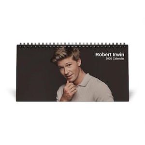 Op de afbeelding: Een bureaukalender met een portret van Robert Irwin. De kalender heeft een zwarte spiraalbinding en toont de tekst "Robert Irwin 2026 Calendar" in het wit. De achtergrond is donkergrijs.