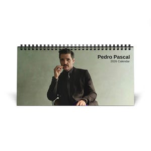 Peut inclure: Calendrier de bureau avec une photo de Pedro Pascal. Le calendrier a une reliure à spirale noire et le texte "Pedro Pascal 2026 Calendar" en blanc. L'image montre Pascal en costume sombre, fond neutre.