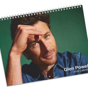 Op de afbeelding: Een spiraalgebonden kalender met een portret van Glen Powell. De afbeelding toont Powell in een spijkeroverhemd tegen een groene achtergrond. De tekst "Glen Powell 2026 Calendar" staat rechtsonder. De kalender is wit en heeft een zilveren spiraalbinding.