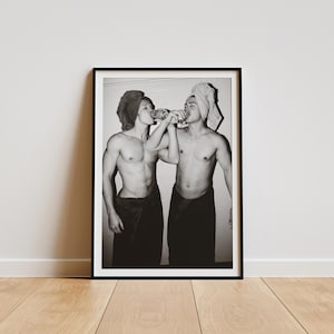 Op de afbeelding: Een zwart-wit ingelijste print met twee personen met ontbloot bovenlijf en handdoeken om hun middel en hoofd gewikkeld. Ze staan naast elkaar en houden elk een glazen fles aan hun lippen alsof ze drinken, vastgelegd in een spontane stijl.