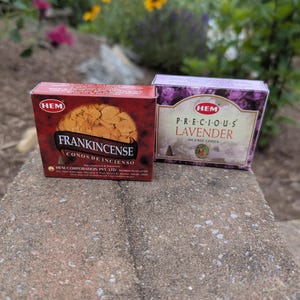 May include: Two boxes of HEM incense cones. One box is red with "FRANKINCENSE" printed on it. The other box is purple with "PRECIOUS LAVENDER" printed on it. The boxes are on a stone surface, with flowers in the background.
