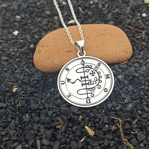 May include: A silver-colored pendant necklace featuring a circular charm with intricate symbols and text. The pendant hangs from a delicate chain and rests on a small, brown stone. The background is dark.