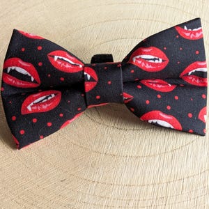 May include: A black bow tie featuring a pattern of red vampire lips and small red dots. The fabric bow tie has a hook and loop fastening. The bow tie is suitable for formal occasions.