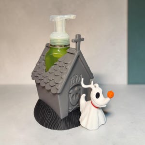 Puede incluir: Un dispensador de jabón gris con forma de caseta de perro, techo de tejas y la palabra ZERO grabada en el frente. Una bomba transparente contiene líquido verde en su interior. Un perro fantasma blanco con collar rojo y nariz naranja está al lado.
