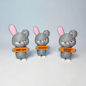May include: Three gray rabbit figurines with pink inner ears and white feet. Each rabbit has a grumpy expression and holds an orange rectangular sign. The signs feature black text reading HOP OFF, BUNARCHY, and HOPING CHAOS respectively.