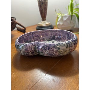 May include: A decorative, oval-shaped bowl with a marbled pattern in shades of purple, white, and green. The bowl has a glossy finish and a unique, double-lobed design. The bowl is a decorative item.