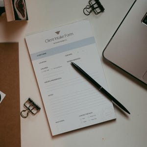 May include: A client intake form with the text "Client Intake Form" and a pen on a white surface. The form includes fields for client name, location, and desired services. A laptop and office supplies are also visible.