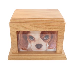 May include: A wooden pet urn with a photo insert featuring a dog's face. The urn is light brown and has a simple design.