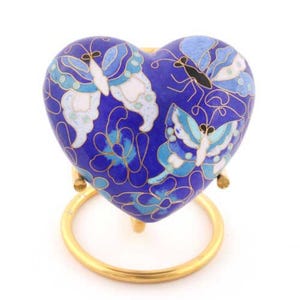 May include: A heart-shaped, blue decorative urn with butterfly and floral designs. The urn is supported by a gold-colored metal stand. The butterflies are white, light blue, and black.