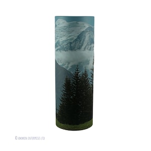May include: A cylindrical cremation urn featuring a mountain and forest scene. The urn depicts a blue sky, snow-capped mountains, and dark green pine trees.