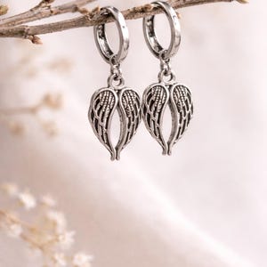May include: A pair of silver-colored hoop earrings with wing-shaped charms. The earrings are suspended from a thin, brown twig against a soft, blurred background. The wing charms have detailed feather patterns.