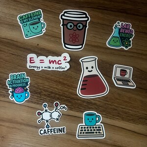 May include: A collection of colorful stickers with coffee and science themes. Designs include a coffee cup, a beaker, a laptop, and the caffeine molecule. Text includes "Caffeine is my element", "Brain activation in progress", and "Energy = milk x coffee".