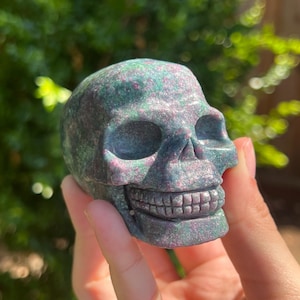 May include: A carved skull made of green and pink speckled stone. The skull has detailed features, including eye sockets, a nose cavity, and a full set of teeth. The skull is held in a hand against a blurred green background.