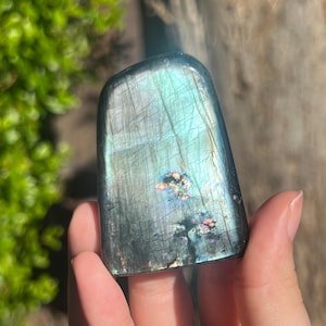 May include: A polished, rectangular labradorite stone with iridescent blue, green, and gold flashes. The stone is held in a hand, showcasing its smooth, reflective surface and natural patterns. The background is blurred, with green foliage.