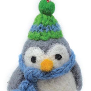 May include: A gray and white felt penguin wearing a green and blue knit hat and a blue scarf. The penguin has black eyes and an orange beak.